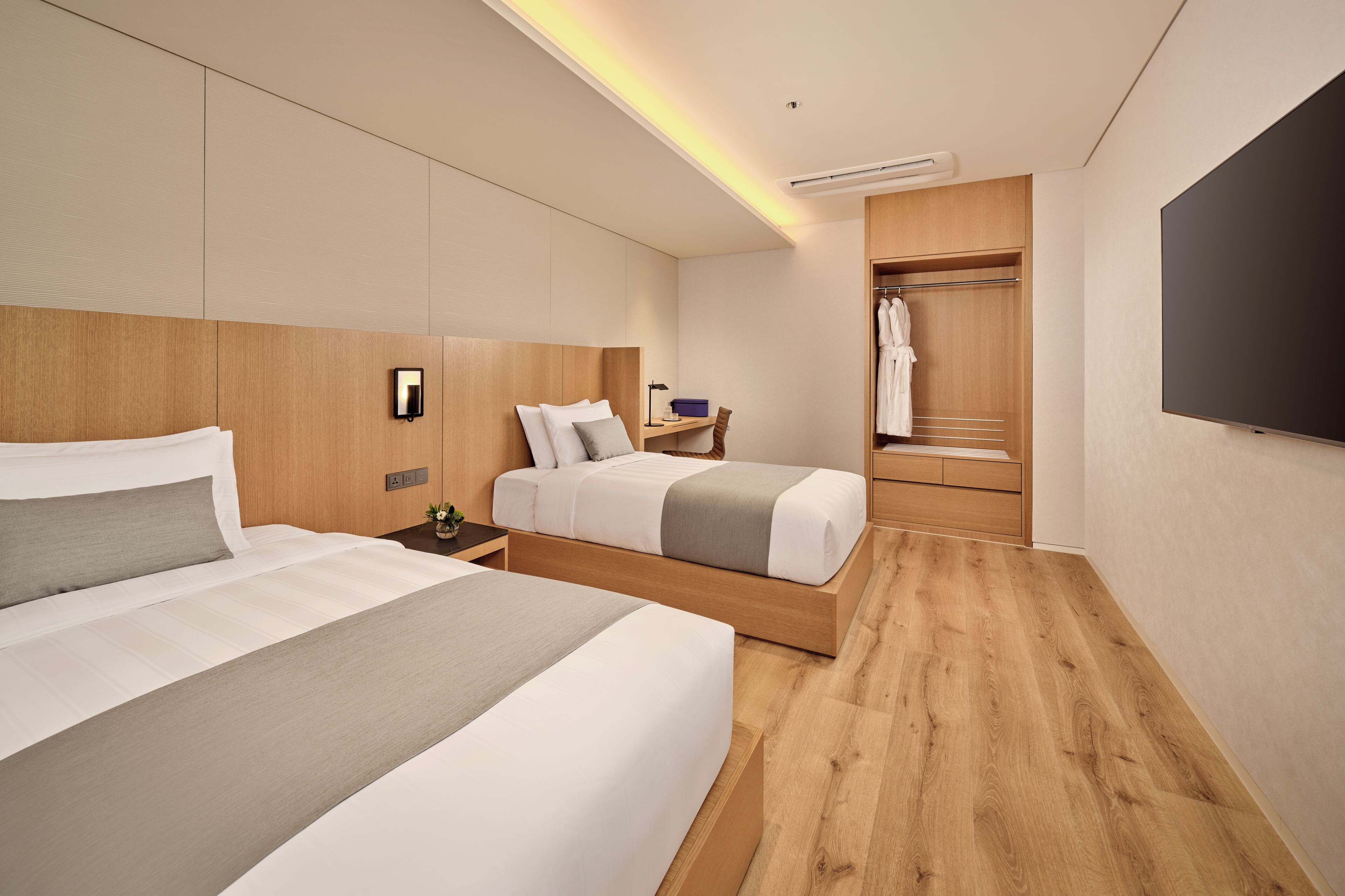 A modern hotel room with two beds, a walk-in closet, a flat-screen TV, and a bedside table with a lamp.