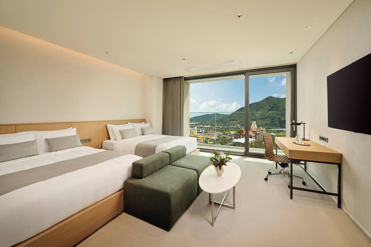 A hotel room with a large bed, a desk, a chair, and a view of a cityscape through a large window.