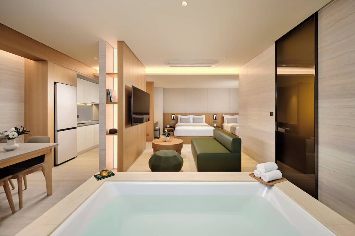 A modern hotel room with a large bathtub, a sofa, a dining area, and a separate bedroom with a bed and bedside tables.