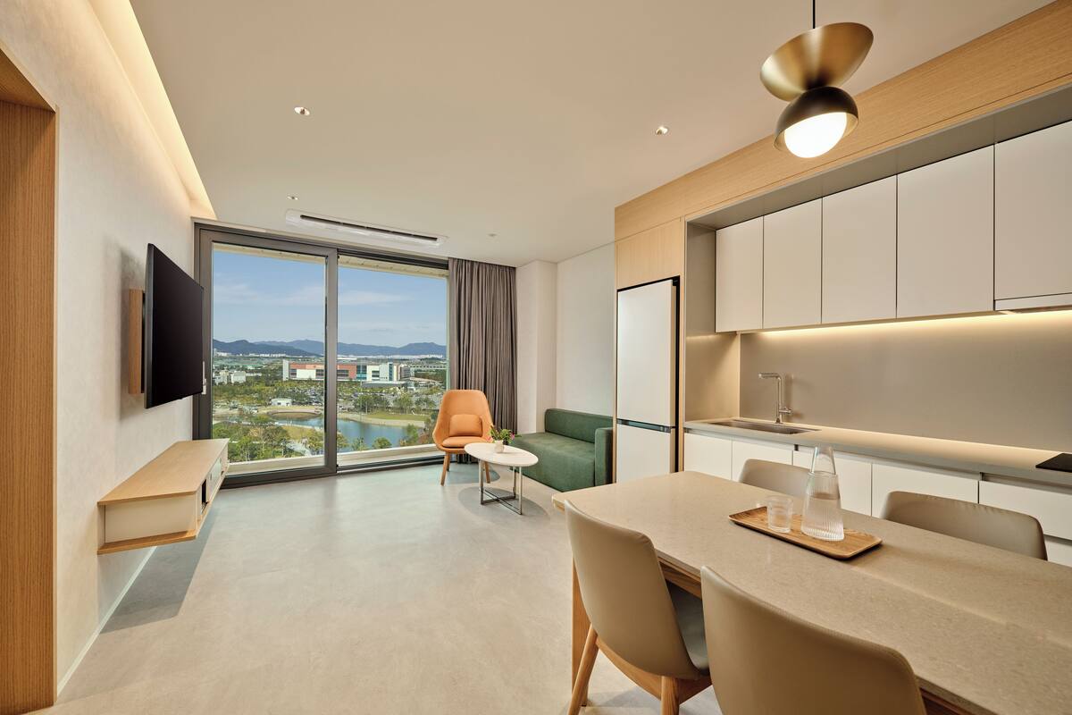 A modern hotel room with a dining area, a sofa, a TV, and a large window offering a city view.