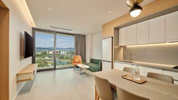 A modern hotel room with a dining area, a sofa, a TV, and a large window offering a city view.