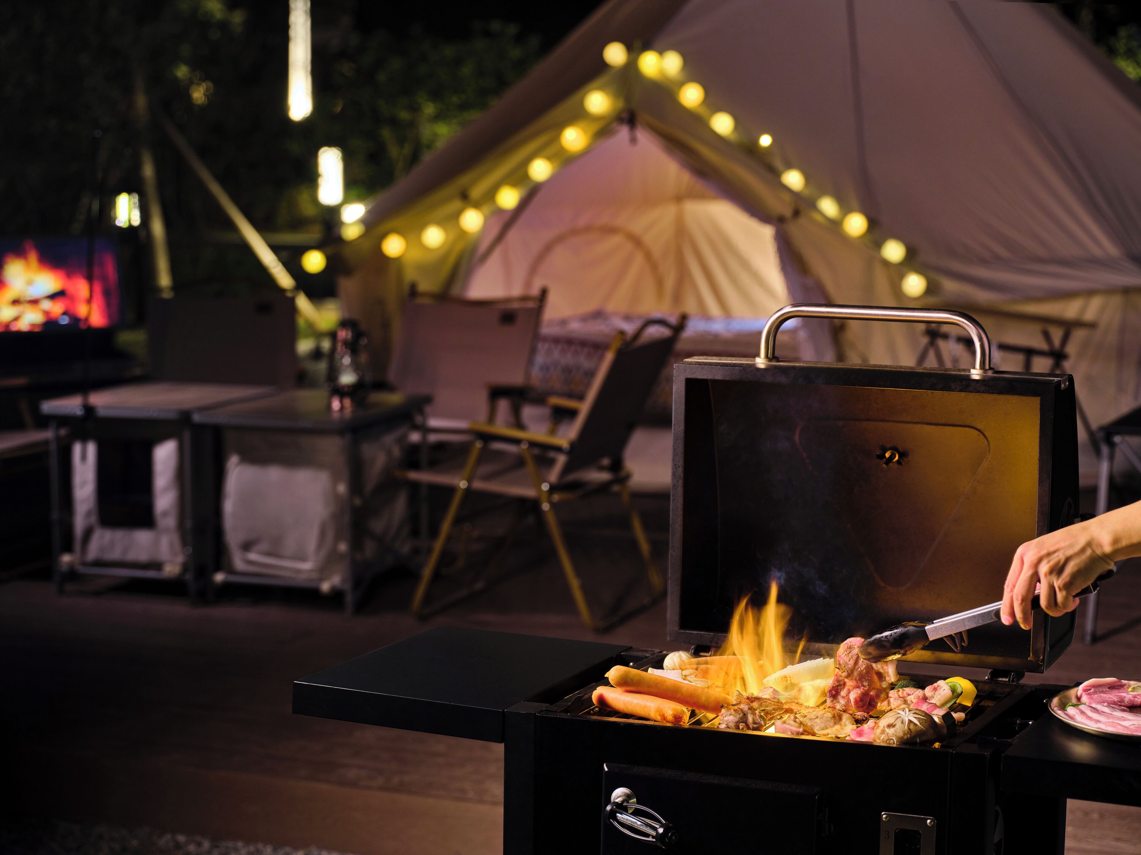 A portable grill with food being cooked, a tent with string lights, and a table with chairs in the background.