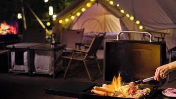 A portable grill with food being cooked, a tent with string lights, and a table with chairs in the background.