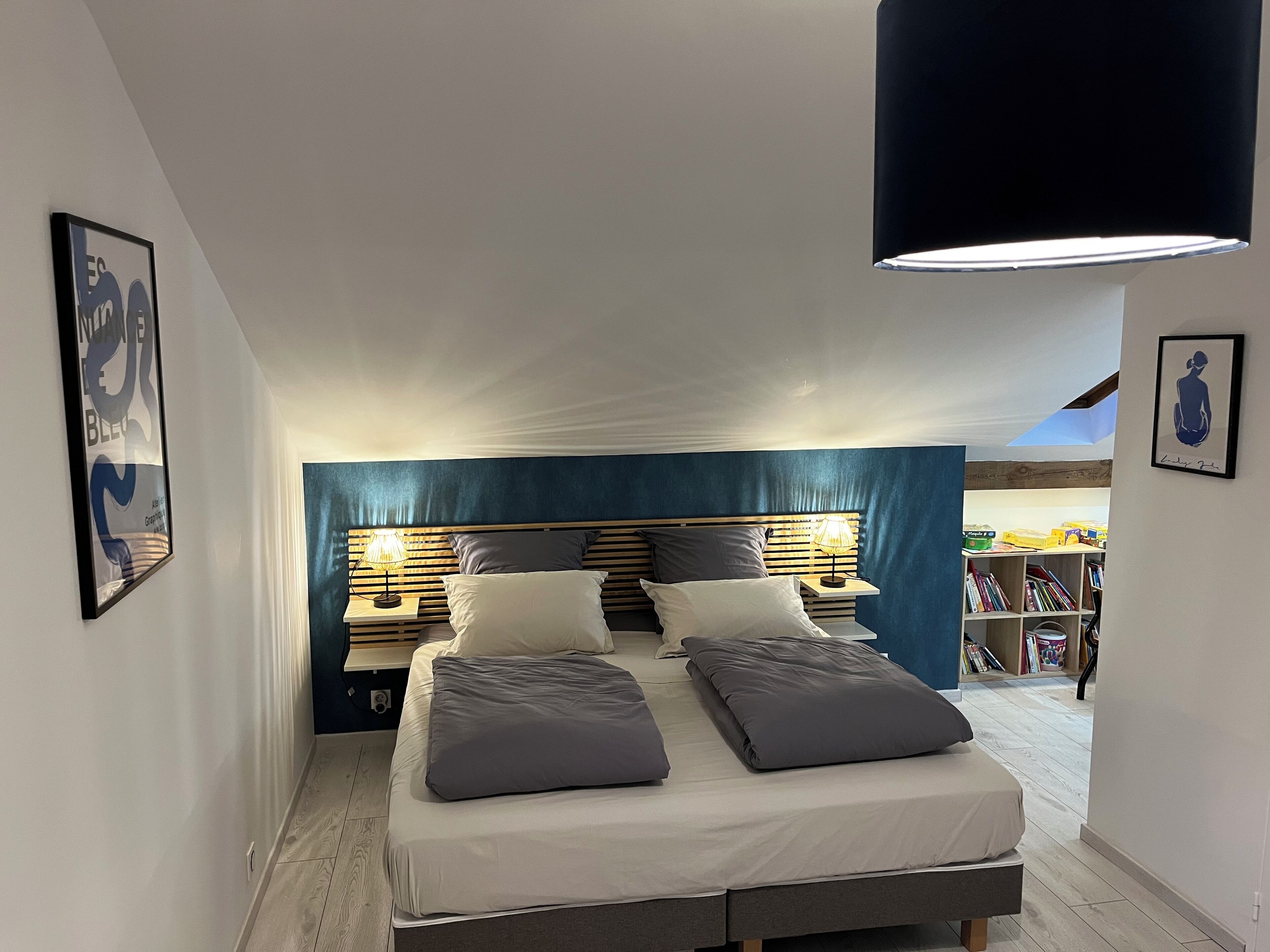 Ein Schlafzimmer mit einem Bett, Nachttischlampen und einer Deckenleuchte. Im Hintergrund ist ein Bücherregal zu sehen.