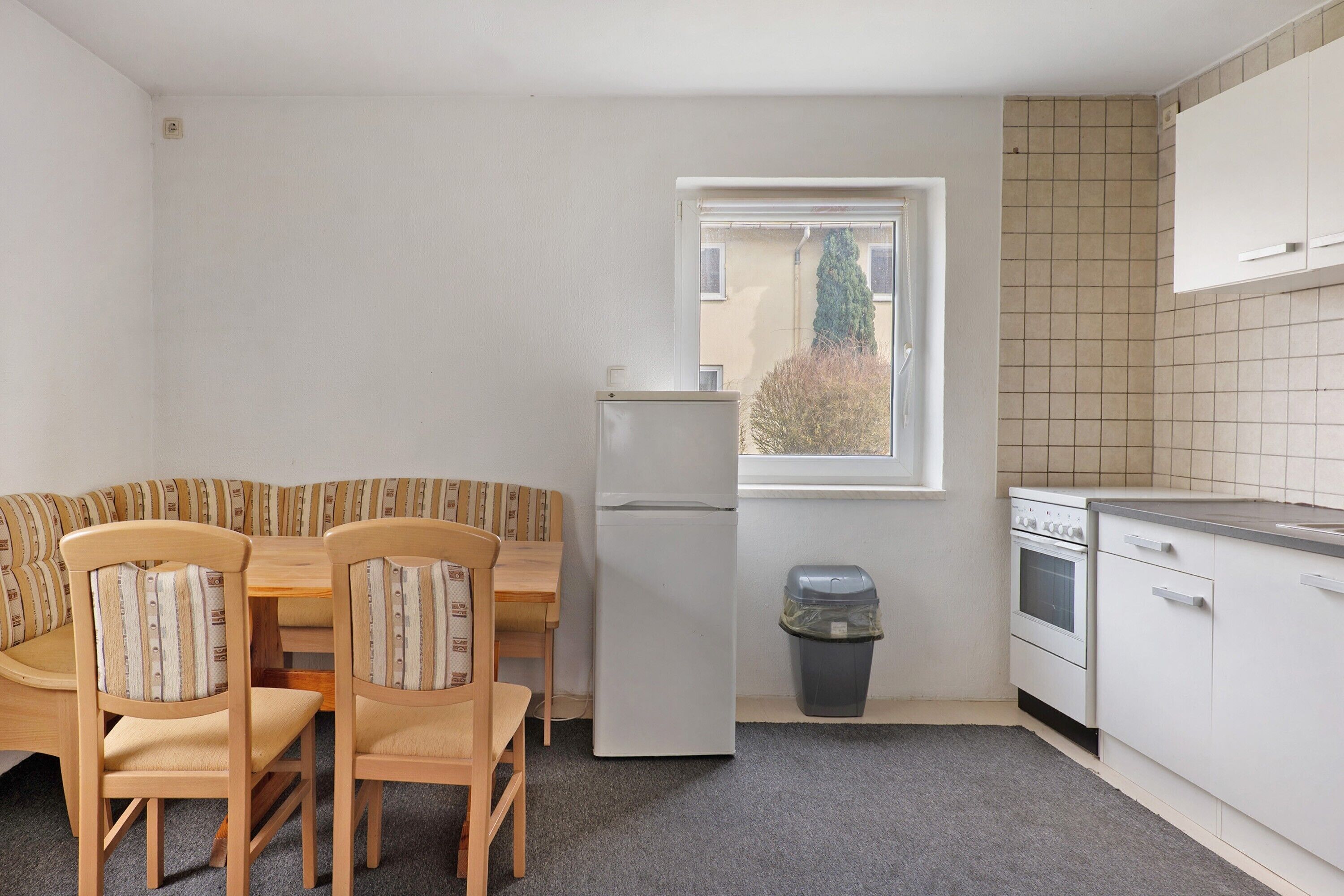 Een kleine keuken met een ingebouwde oven, een koelkast en een raam met uitzicht op bomen.