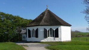 Une petite église blanche, avec un toit de chaume et une croix au sommet, entourée de verdure.