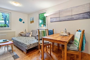 A cozy room with a bed, a dining table, and chairs. There are windows with views of trees and a lamp on the table.
