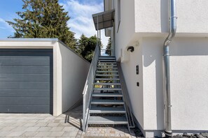 A modern exterior with a metal staircase leading to a covered entrance, a garage door, and a white wall with a metal pipe.