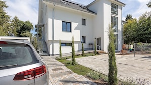 A modern two-story house with a car parked in the driveway, a staircase leading to the entrance, and a small garden area with young trees.