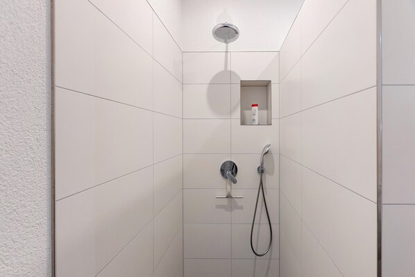 A shower with a handheld showerhead and a handheld faucet, a small shelf with a bottle, and tiled walls.