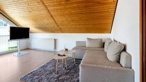 A modern living room with a wooden ceiling, a flat-screen TV, a grey sofa, and a small round table with a remote control. - Apartment 'Fewo Georg' with Wi-Fi (Pfullendorf)