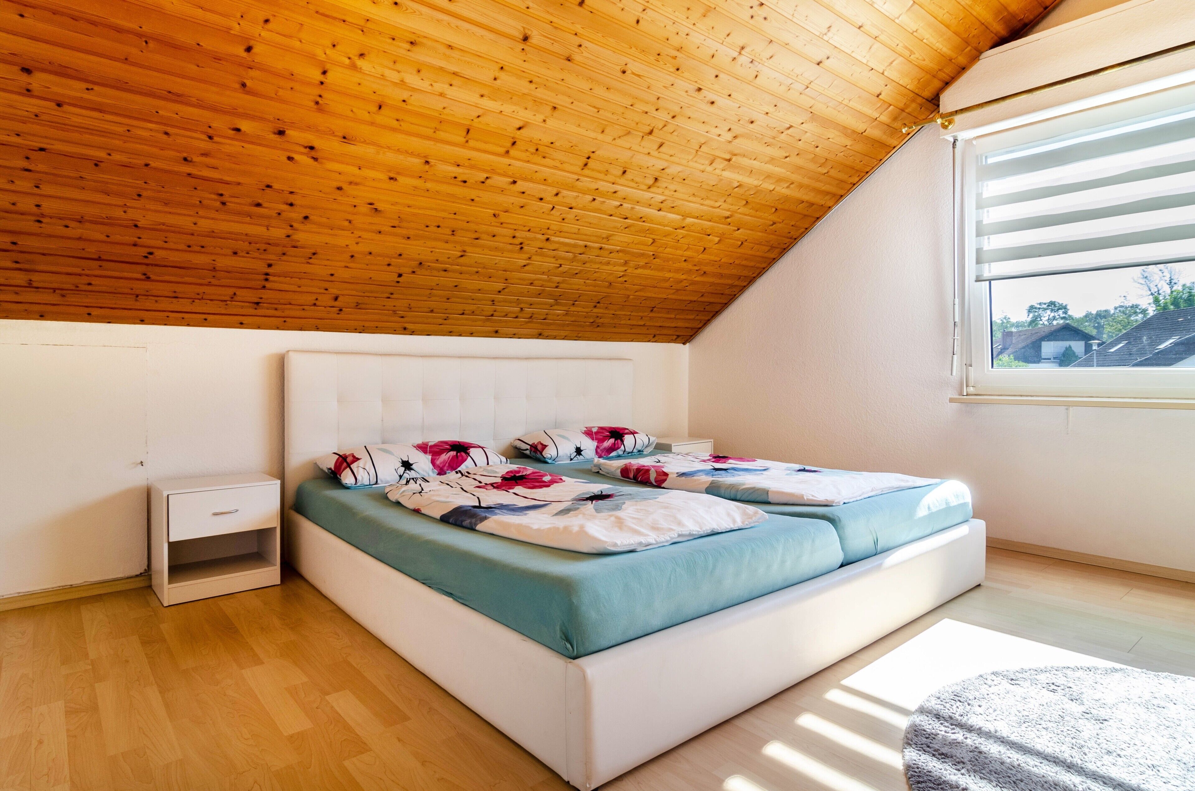 A bedroom with a sloped ceiling, a bed with floral bedding, a bedside table, and a window with blinds.