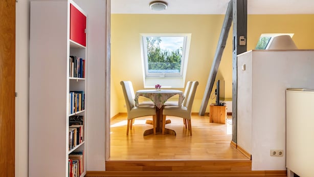 Ein Zimmer mit Holzboden, einem Esstisch mit Stühlen, einem Bücherregal und einem Fenster mit Blick ins Grüne.