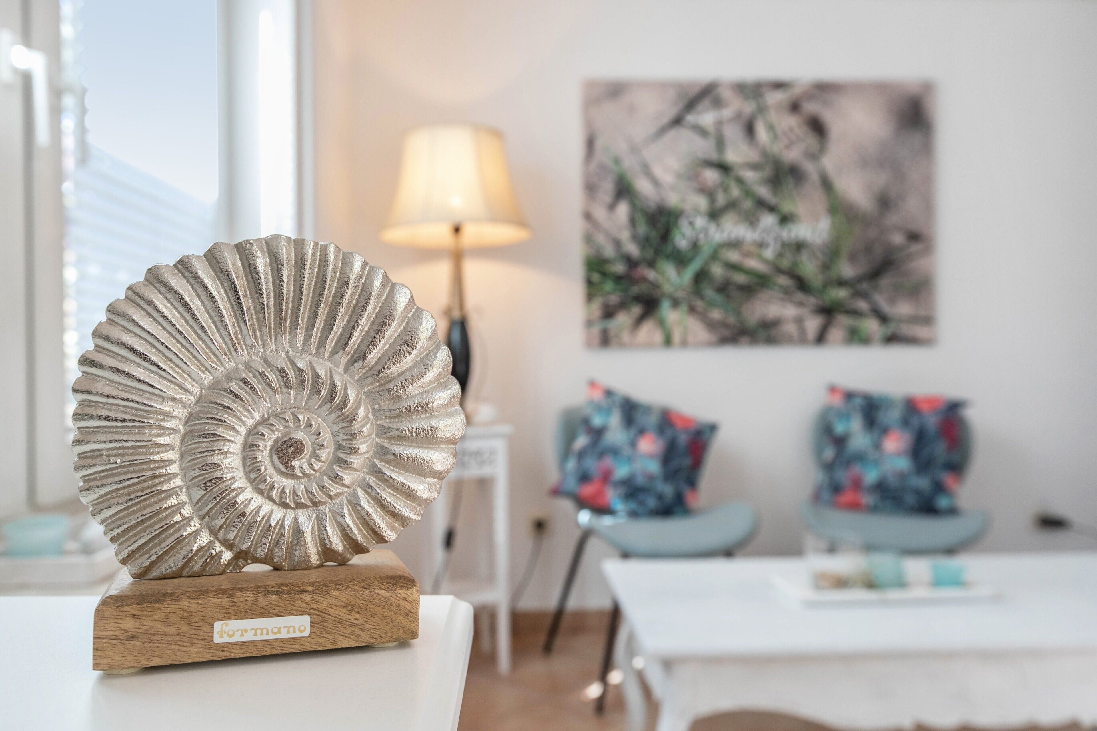 Una stanza con una conchiglia decorativa su un supporto di legno, una lampada, un quadro e due poltrone con cuscini floreali.