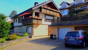 Un immeuble résidentiel de deux étages, doté d’un garage, d’un balcon orné de fleurs et d’une voiture garée.