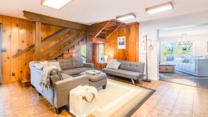 A spacious living room with a large sofa, a smaller sofa, and a coffee table. There is a staircase with wooden railings leading to an upper level. The room has wooden walls and a large window providing natural light.