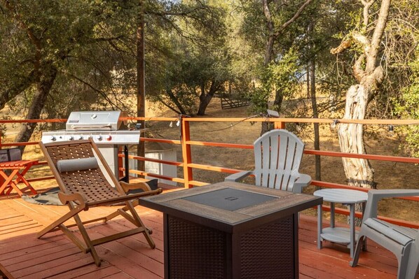 A deck with outdoor furniture, including a grill and chairs, surrounded by trees. - Big Backyard with games, hot tub, Firepit, Deck, Views (Oakhurst)
