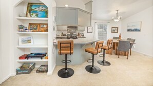 Cozinha moderna com armários brancos, uma ilha central com banquetas e uma área de jantar com mesa e cadeiras de madeira.