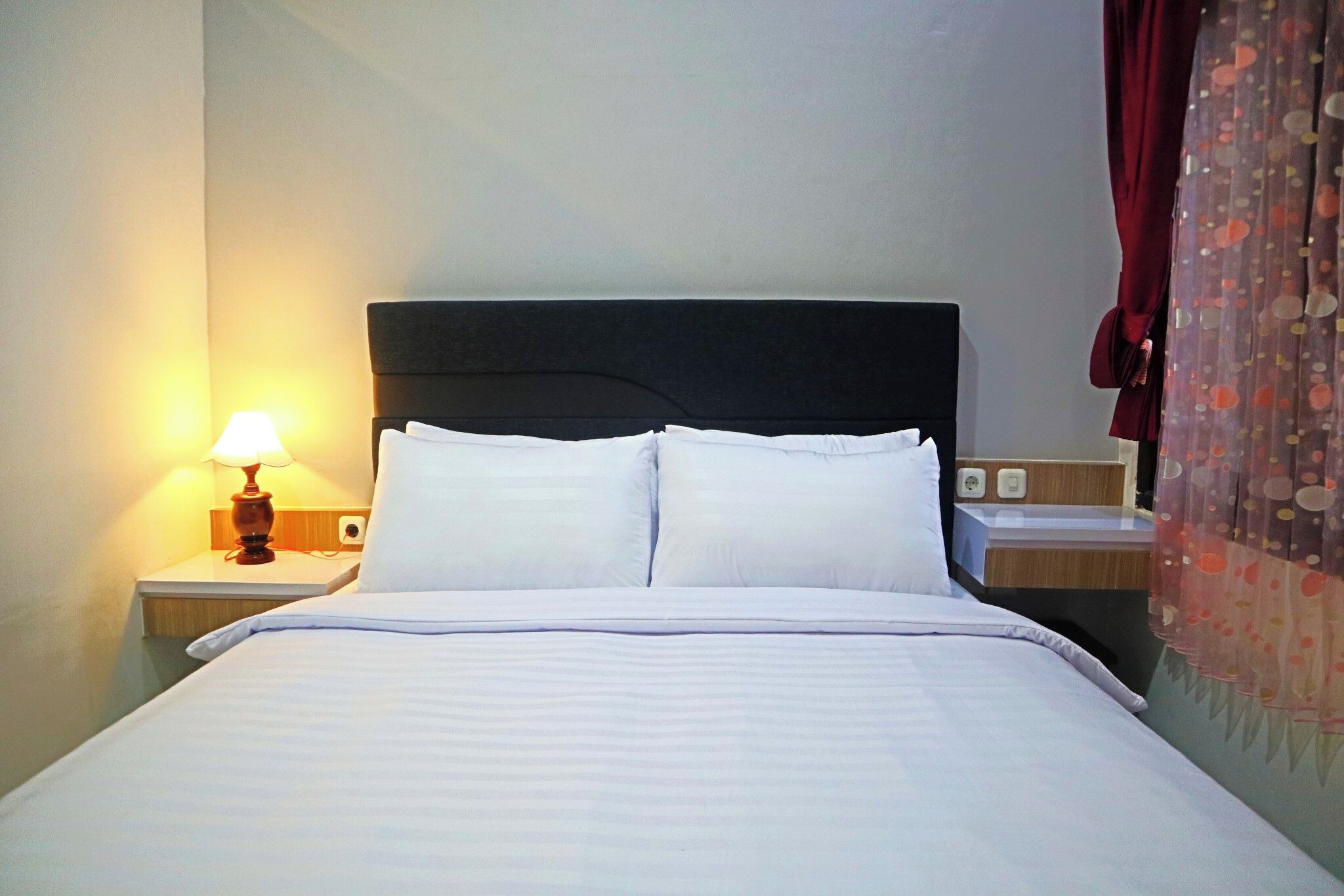 Una cama bien tendida con sábanas blancas, una lámpara de noche y un enchufe en la mesita de noche.