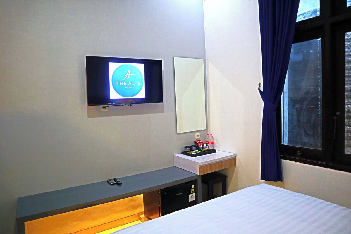 A hotel room with a bed, a desk with a computer, a TV mounted on the wall, a small bedside table with a bottle and snacks, and a window with curtains.