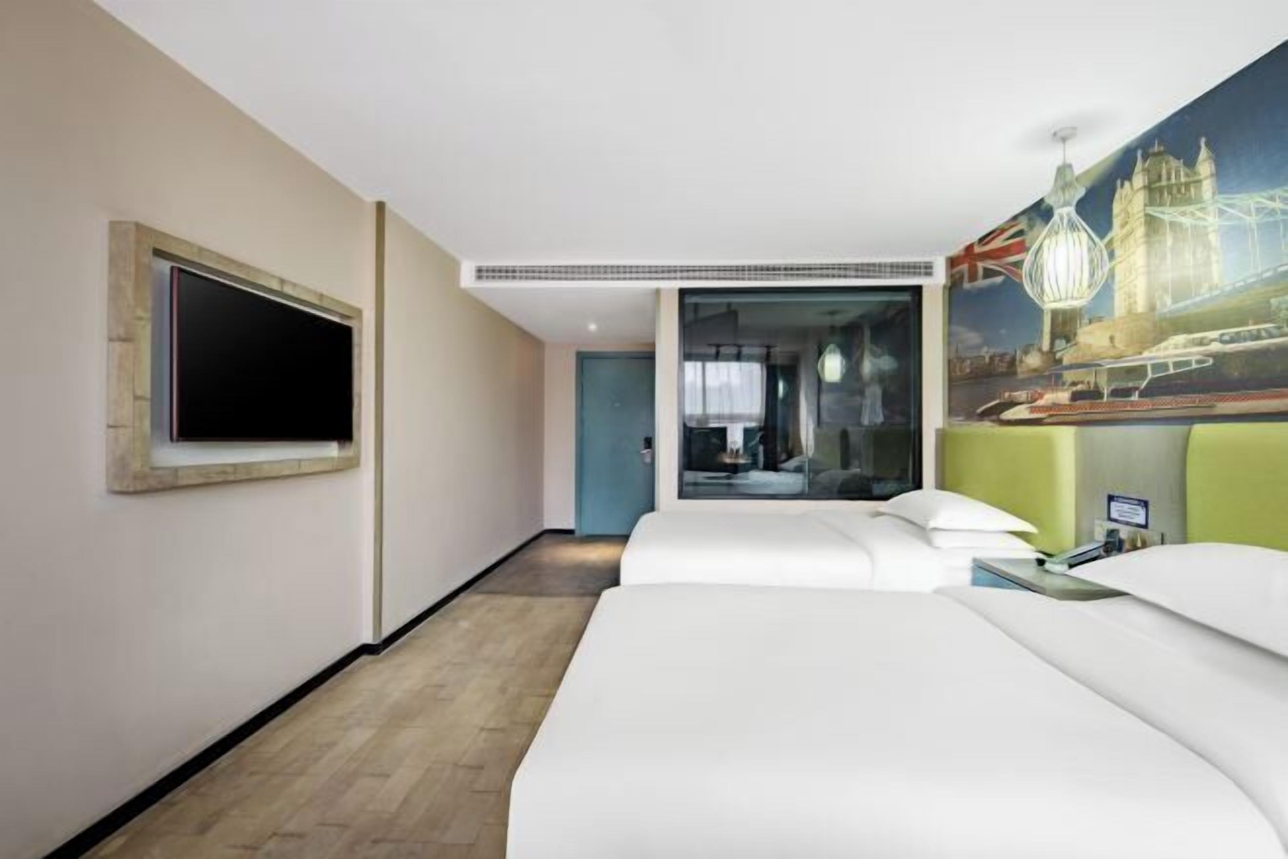Ein Hotelzimmer mit einem großen Bett, einem an der Wand befestigten Flachbildfernseher, einem Stadtbildgemälde und einem grünen, gepolsterten Kopfteil.