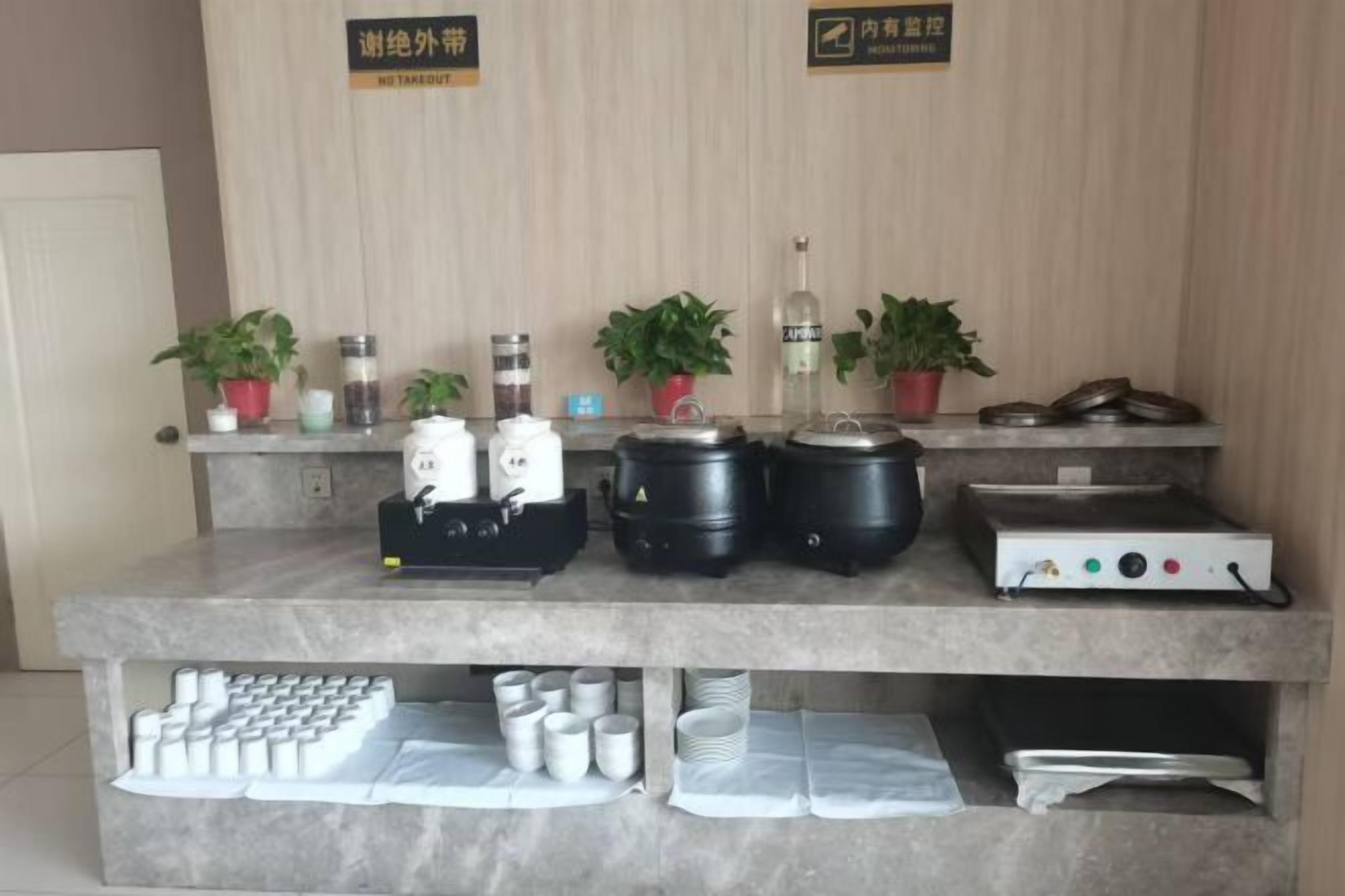A kitchen area with various appliances, including a rice cooker, a pressure cooker, and a rice cooker with a lid. There are also several potted plants and a shelf with jars and containers.