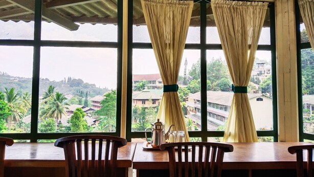 Una habitación con una mesa y sillas de madera, ventanas amplias con cortinas y vista a una zona residencial con casas y vegetación.