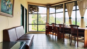 Una habitación con un sofá de cuero marrón, una mesa de vidrio, sillas de madera, una mesa de comedor y ventanas grandes con cortinas.