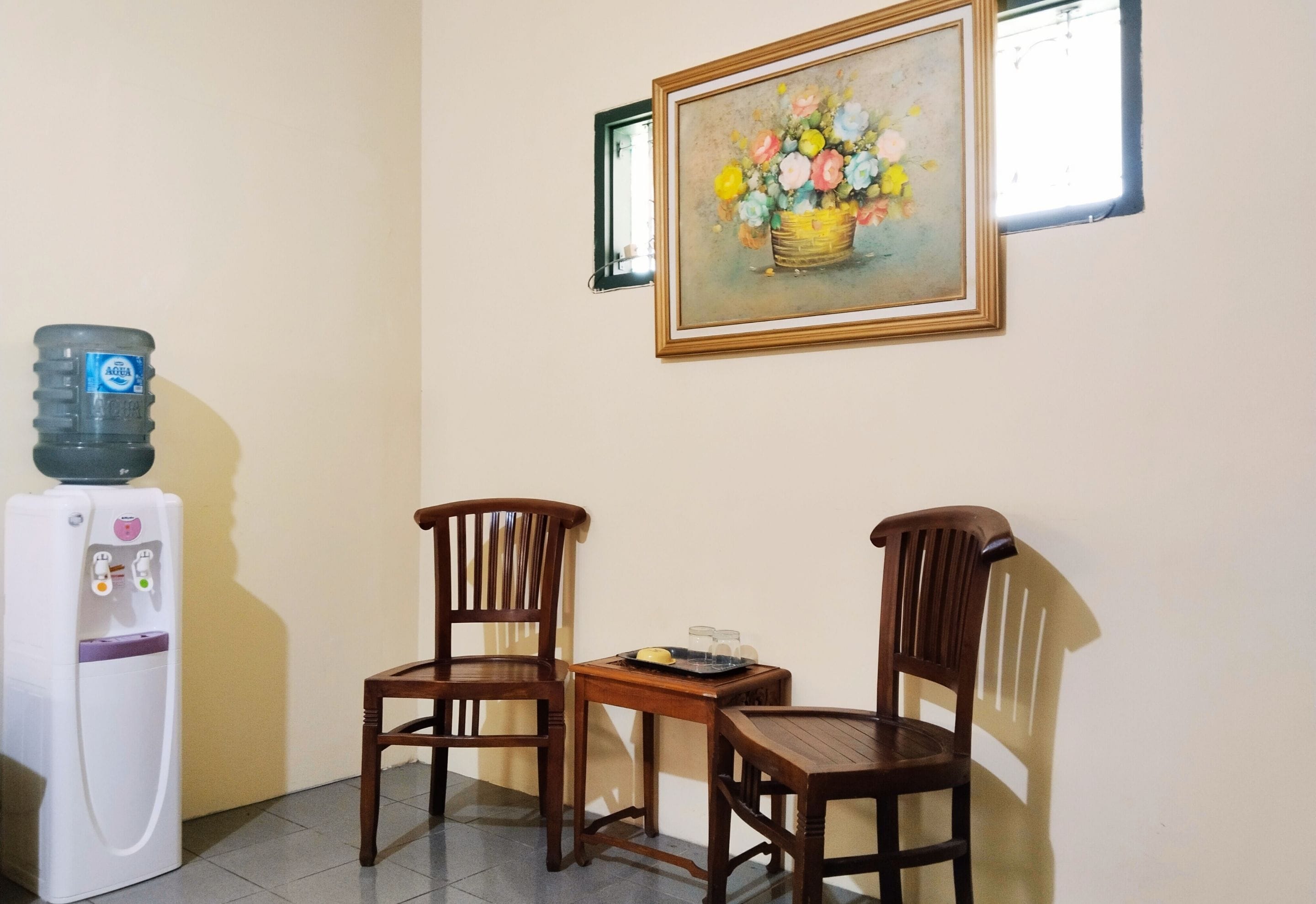 A room with two wooden chairs, a small table, a water dispenser, and a framed painting on the wall.