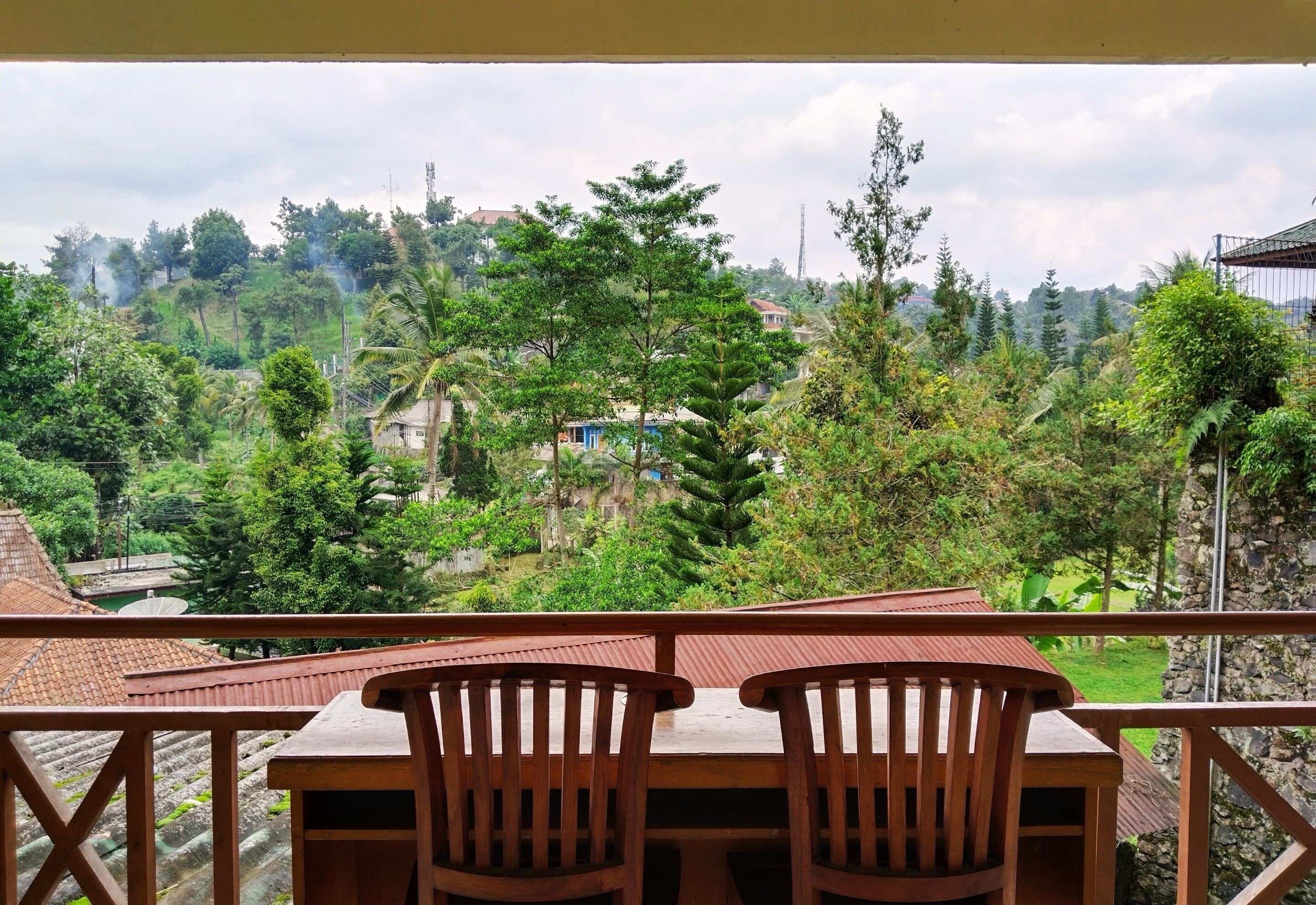 Un balcón con dos sillas de madera y una mesa, con vistas a un exuberante paisaje verde con árboles y algunos edificios a lo lejos.