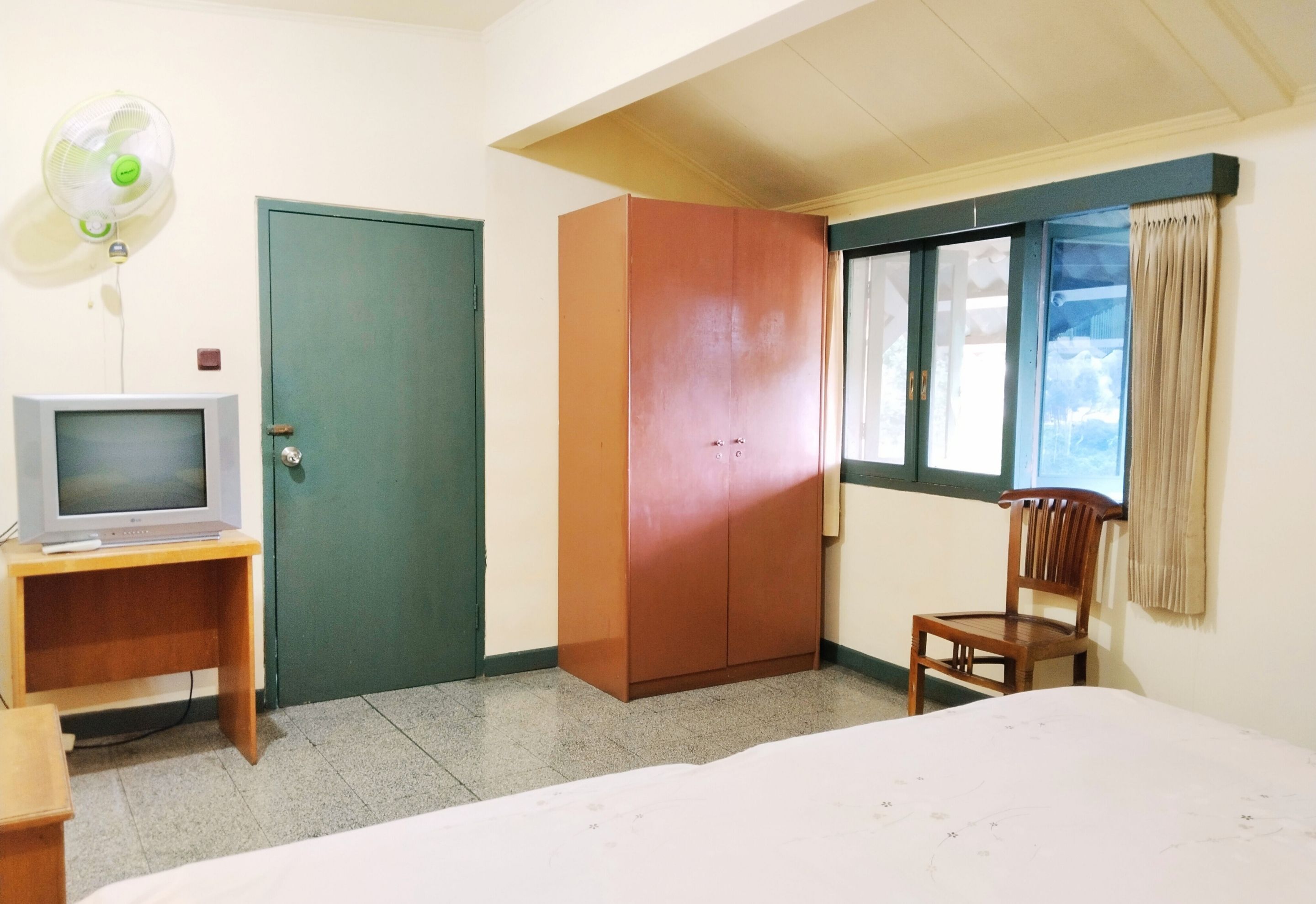 A room with a bed, a wooden chair, a wardrobe, a television on a stand, and a window with curtains.