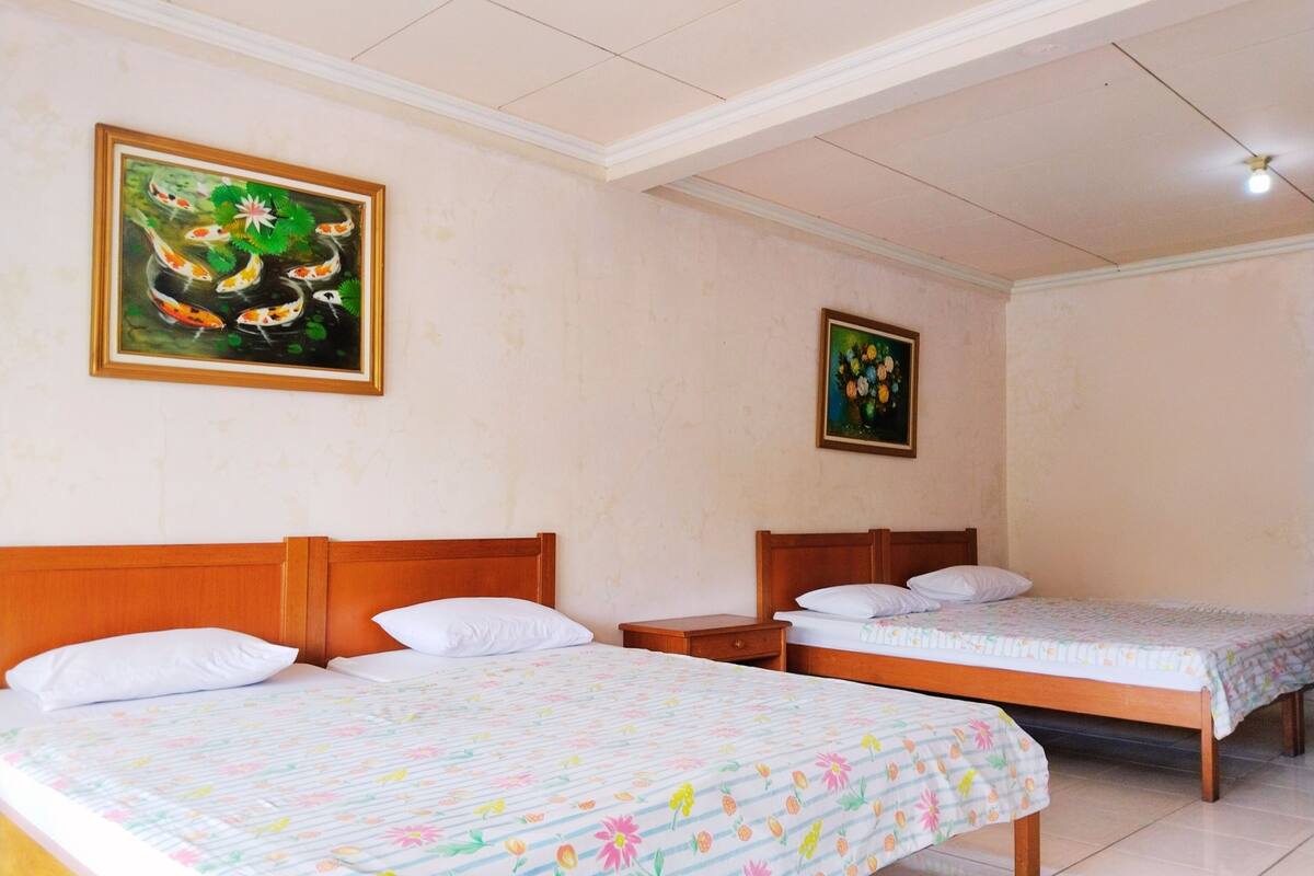 Een hotelkamer met drie bedden, elk voorzien van een bloemenprint beddeken, houten hoofdeinden en witte kussens. Twee ingelijste schilderijen hangen aan de muur, waarvan één met koi karpers en de andere met bloemen.
