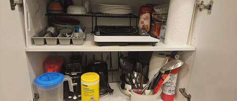 A kitchen cabinet with utensils, a coffee maker, and cleaning supplies.