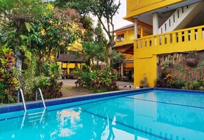 A swimming pool with a blue border, surrounded by lush greenery and a yellow building with a staircase. - Hotel New Sabrina (Cisarua)