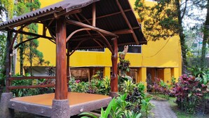 A wooden gazebo with a bench, surrounded by lush tropical plants and a yellow building in the background. - Hotel New Sabrina (Cisarua)