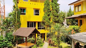 A yellow building with multiple windows, surrounded by lush greenery and a small wooden structure. - Hotel New Sabrina (Cisarua)