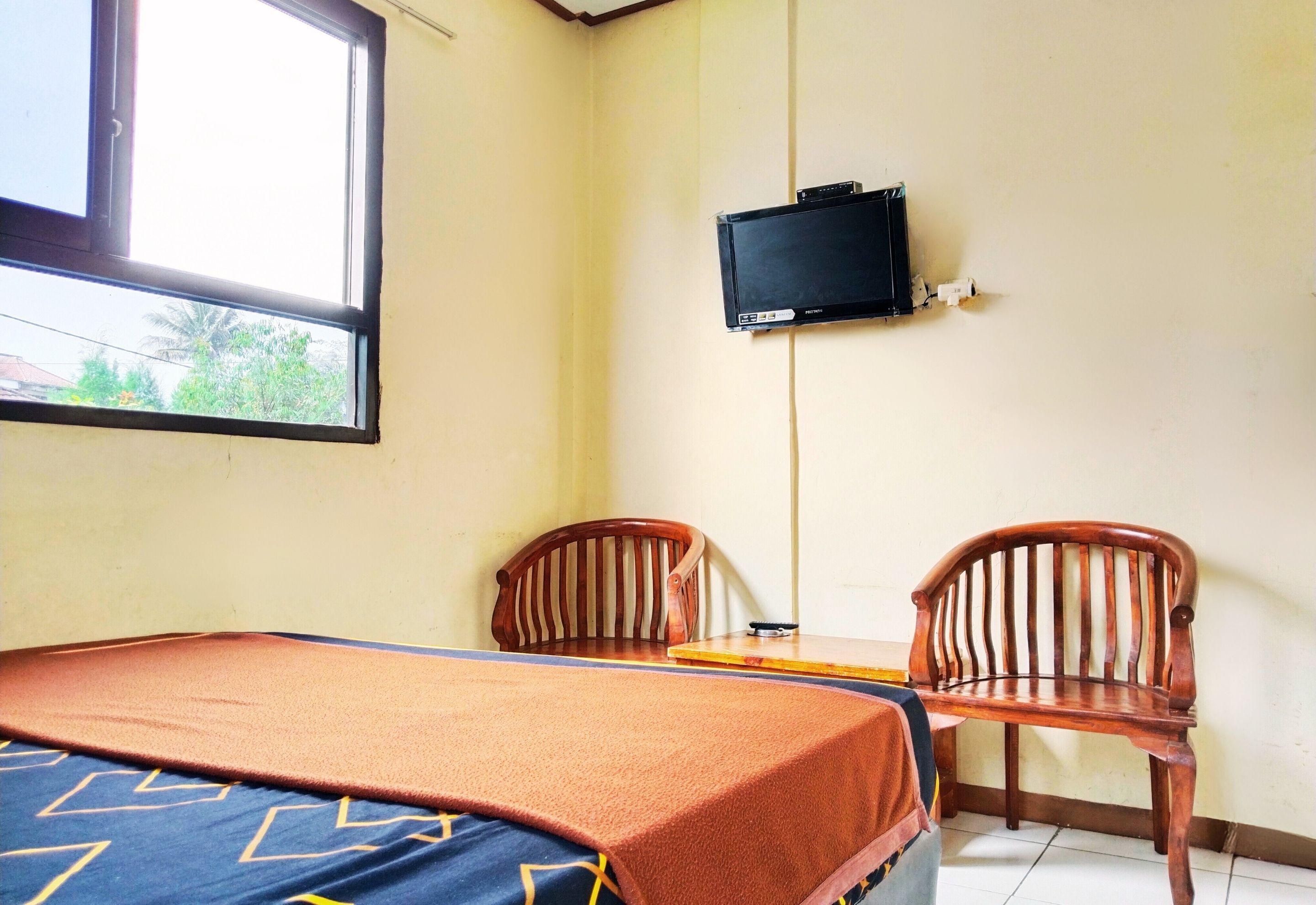 A room with a bed, two wooden chairs, a small table, a television mounted on the wall, and a window.