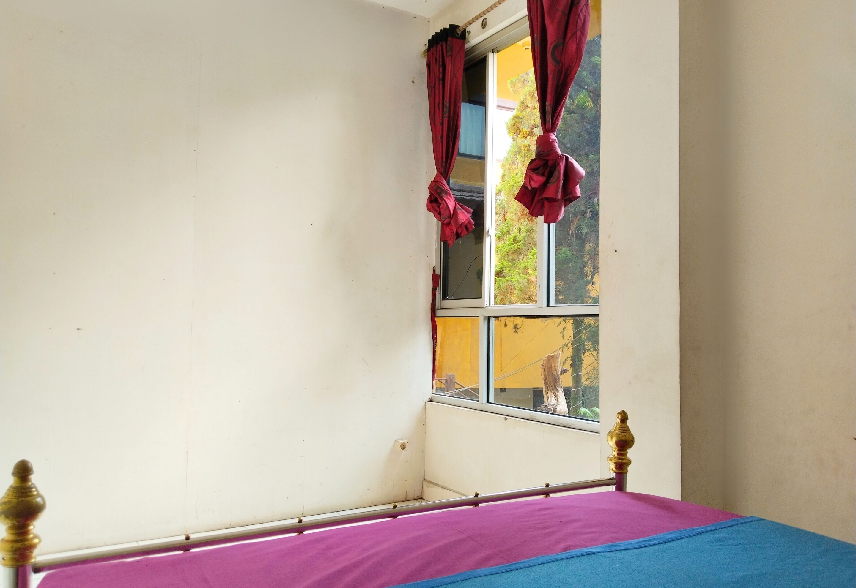 Una habitación con cama, ventana con cortinas y vistas a árboles y un edificio.