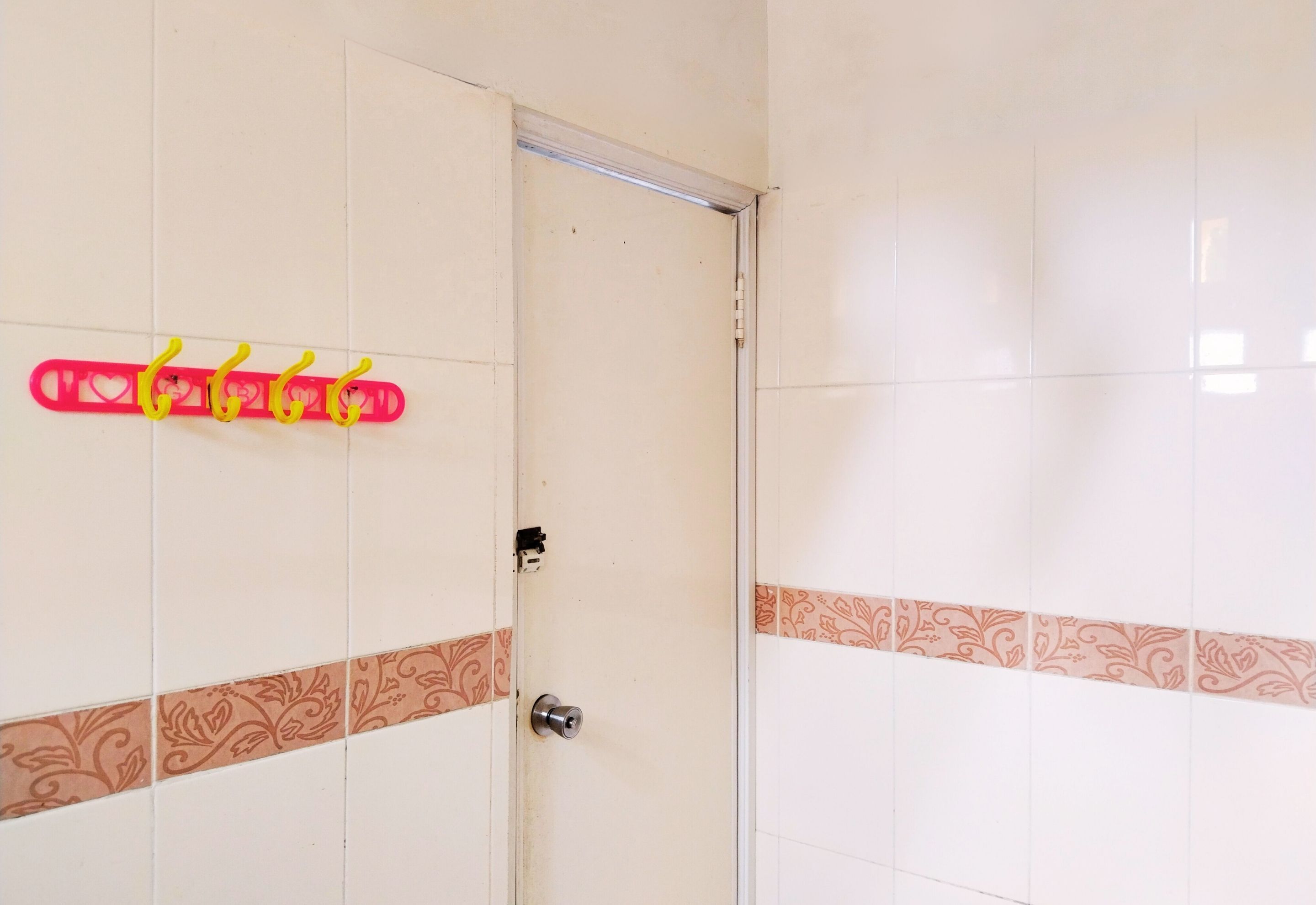 Un baño con azulejos blancos y una puerta con manija. Se observa un toallero rojo con ganchos amarillos.