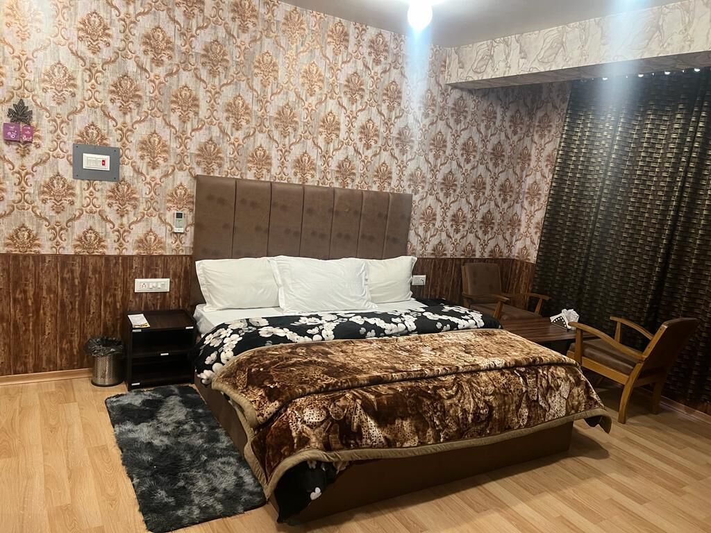 Un dormitorio con una cama grande, cabecera de madera, mesita de noche, una silla y papel tapiz estampado.