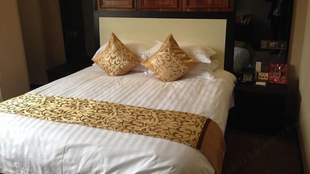 A hotel room with a bed, two decorative pillows, a wooden headboard, and a nightstand with a clock and a bottle.