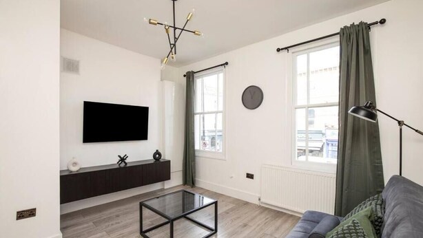 En moderne stue med et fladskærms-tv, et glasbord, en mørk sofa og et stort vindue med gardiner.