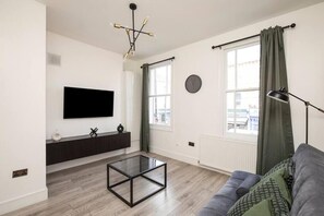 A modern living room with a flat-screen TV, a glass coffee table, a dark sofa, and a large window with curtains. - Spacious Serenity in the Heart of Central London (London)