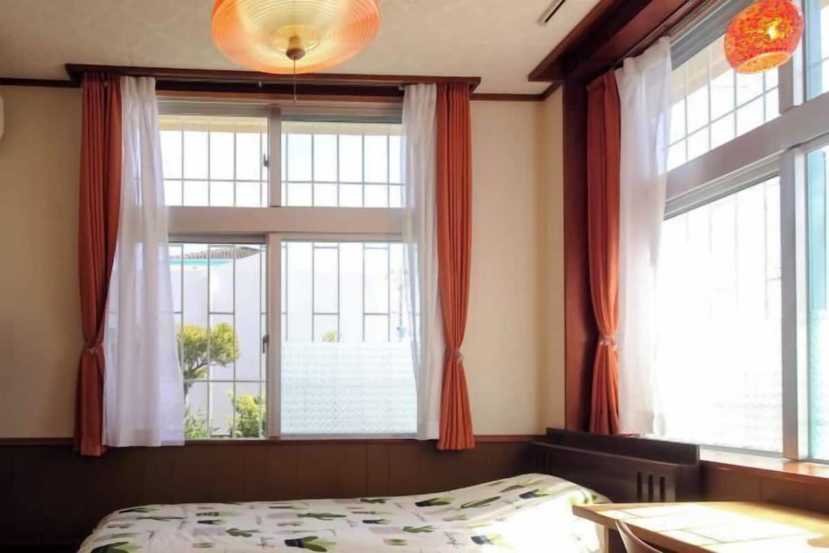 A hotel room with a bed, a table, a window with curtains, and a hanging light fixture.