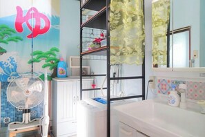 A room with a white cabinet, a white washing machine, a fan, and a shelf with various items. - Minshuku Itoman Bettei Goyukkuri (Itoman)