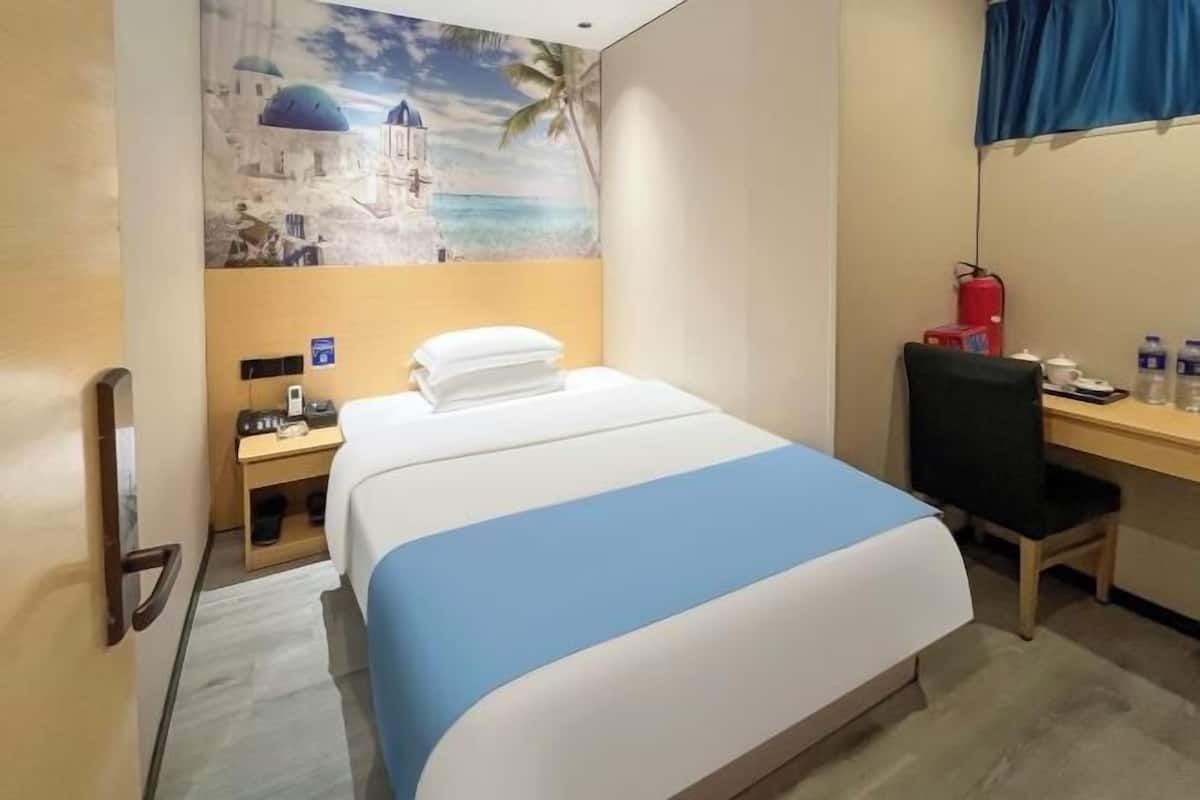 A hotel room with a bed, a desk, a chair, a nightstand, a wall-mounted fire extinguisher, and a painting of a coastal scene.