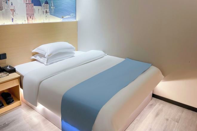 A neatly made bed with white and blue bedding, a bedside table with a lamp, and a window with a view of buildings.