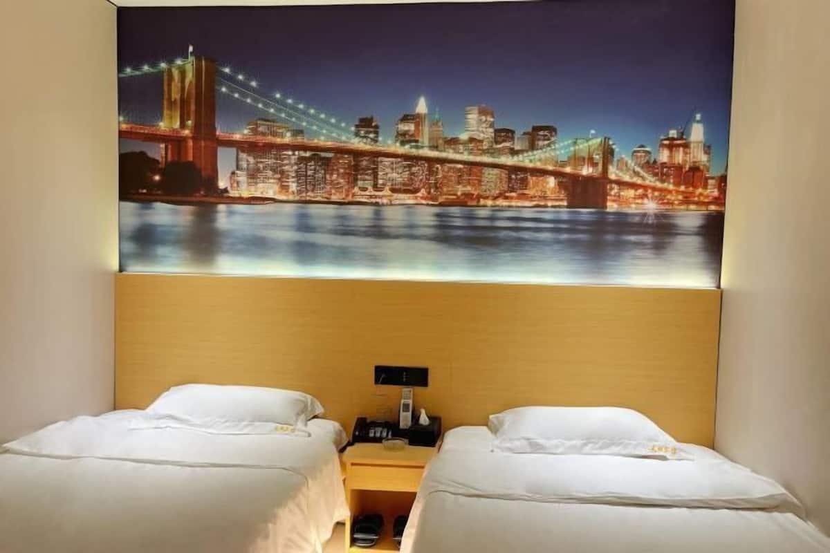A hotel room with two beds, a nightstand with a lamp, and a large cityscape painting on the wall.