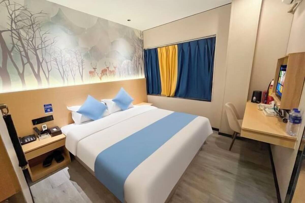 A hotel room with a bed, a desk, a chair, a nightstand, a wall mural, and a window with curtains.