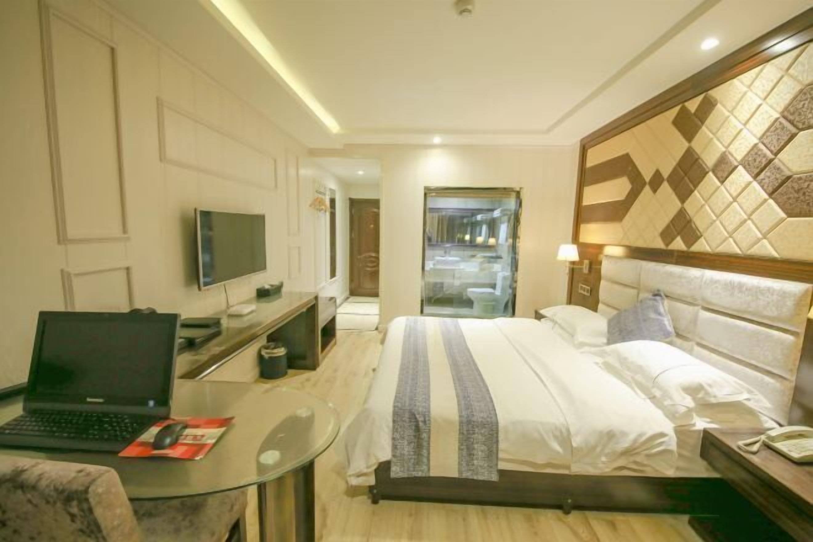 A hotel room with a bed, a desk with a laptop, a television, and a glass table.